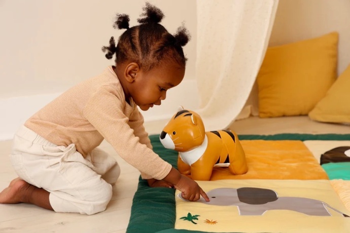 Criança a brincar com um tigre de pelúcia da marca Little Big Friends sobre um tapete colorido.